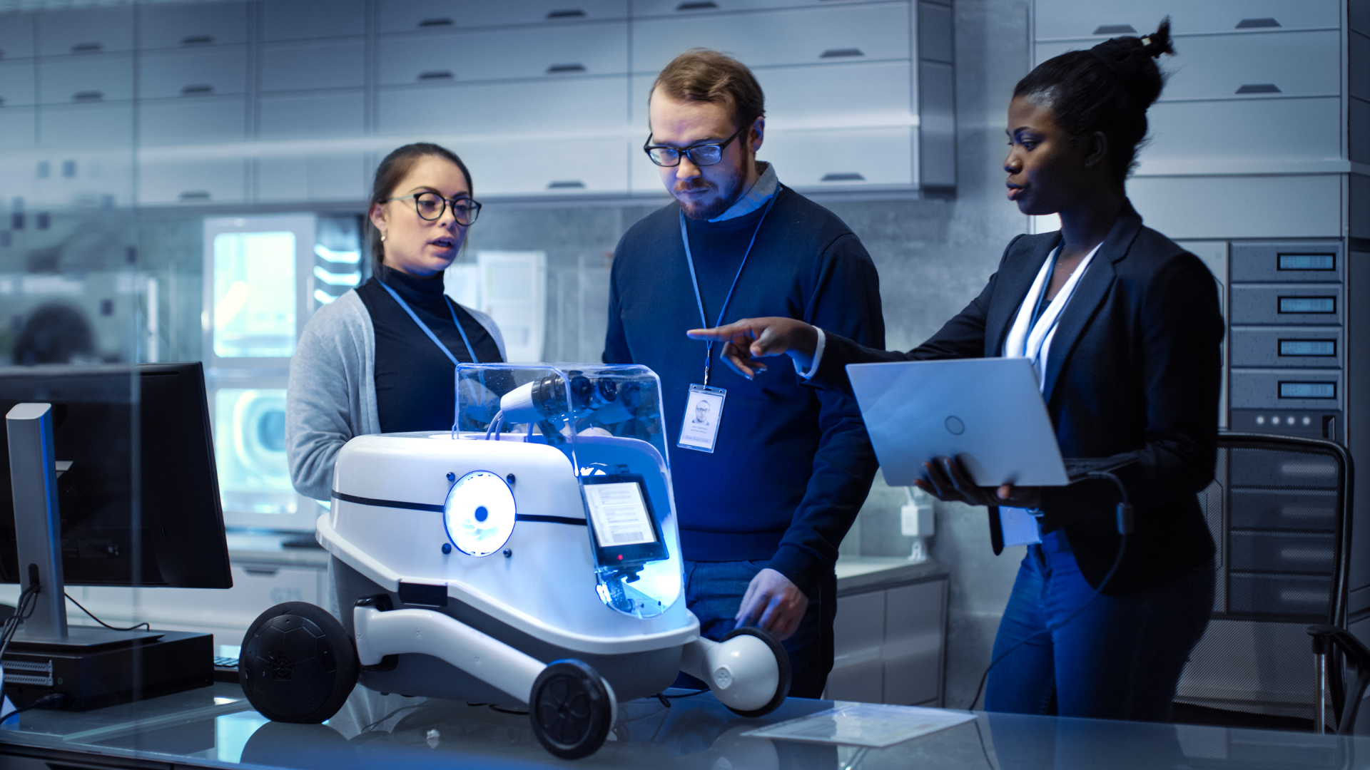 Three engineers wearing lanyards in a sleek modern facility gathered around a small black and white wheeled robot.