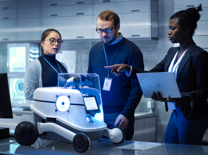 Three engineers wearing lanyards in a sleek modern facility gathered around a small black and white wheeled robot.